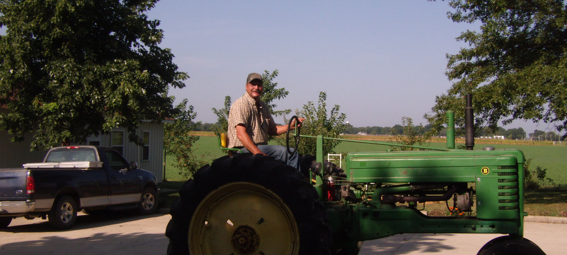 Man in tan plaid shirt and blue jeans on green tractor, blue pickup under tree by house.