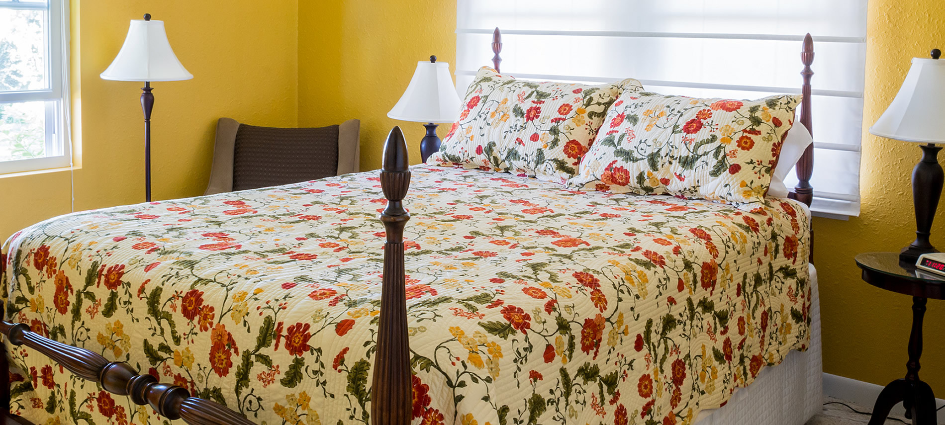 Well-lit gold painted room with lamps all around, floral cream comforter, tan chair in corner.