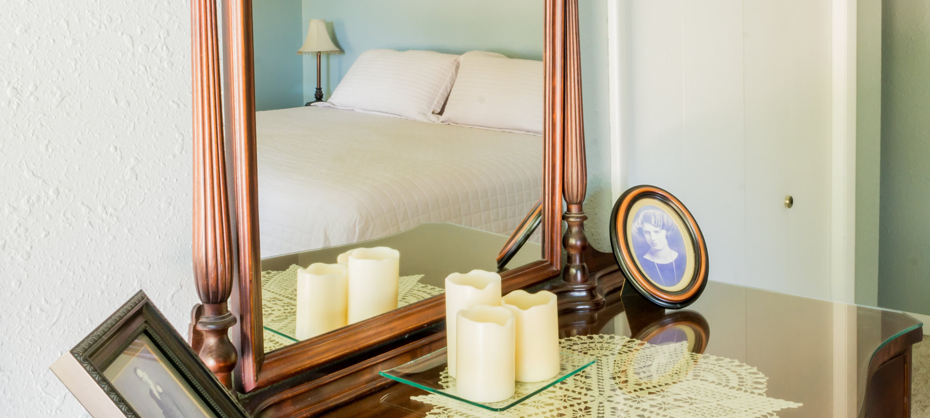 Antique dresser with glass top with reflection of queen bed and cream spread, Candles on glass tray, framed prints.