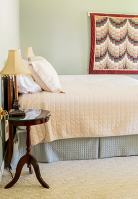 Side view of bed with olive bedskirt and cream spread, cream carpet, olive and cream walls, nightstand with lamp.