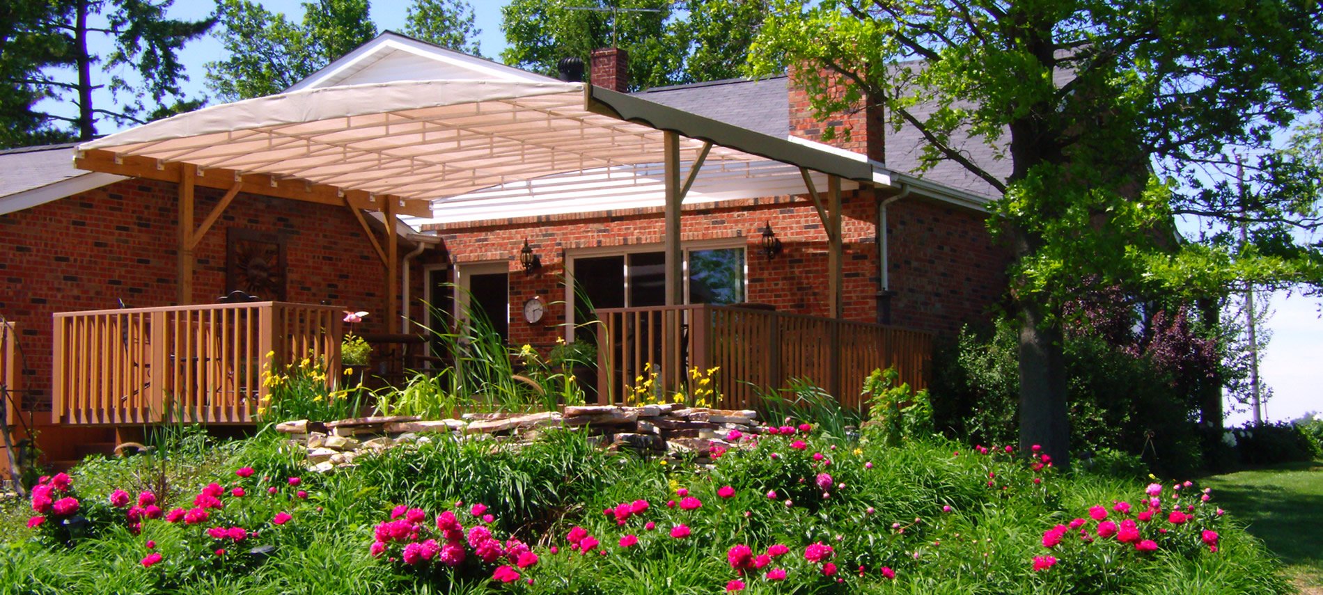 Red-brick house surrounded by trees, pink flowers against stone wall, clock on wall, wood railing.