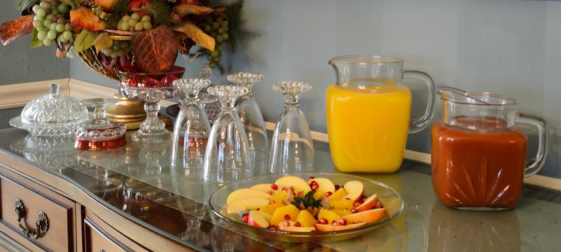 Antique buffet with crystal glasses, juice decanters with tomato and orange juice, fruit bowl with green grapes and apples.