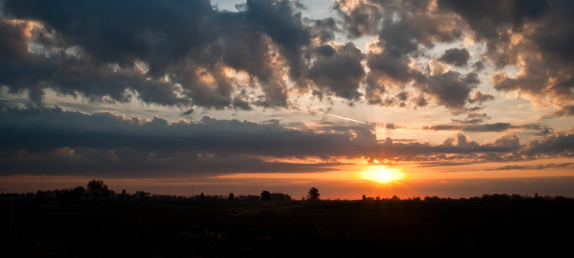 View across horizon at sunset with trees and dark clouds overhead.