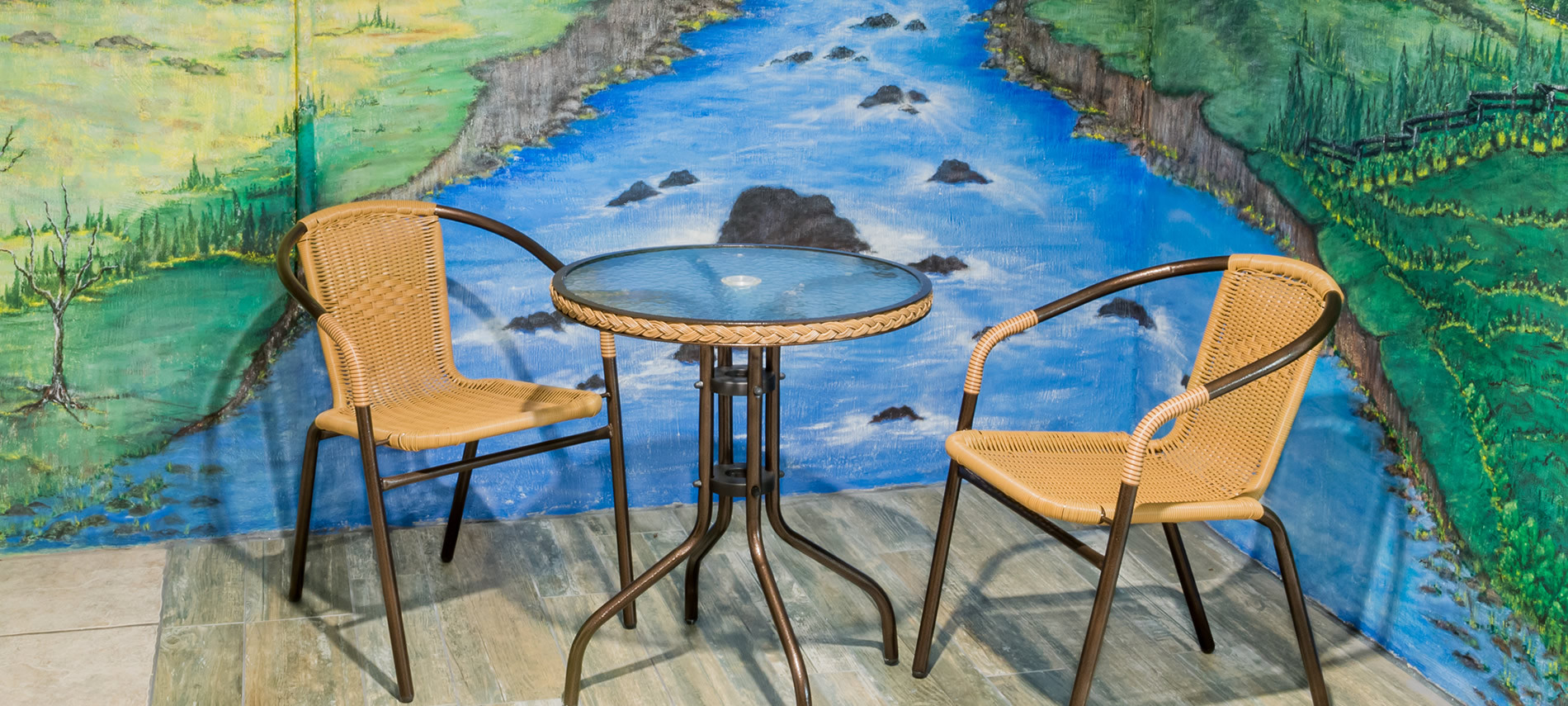 Painting of rocky stream with green grass on sitde, glass table with wicker chairs on wood deck.